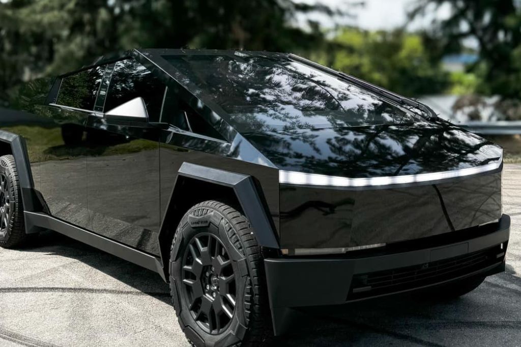 A sleek black Tesla Cybertruck parked outdoors, reflecting trees and sky on its glossy surface. The dark-tinted windows enhance the vehicle's futuristic design while offering privacy and sun protection.