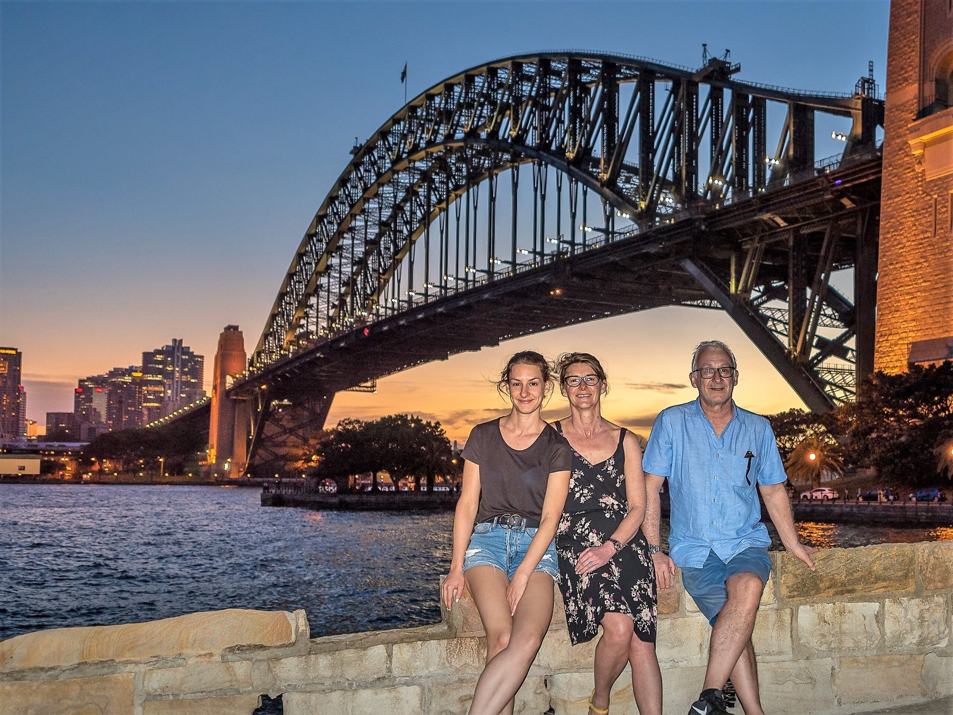 Three people are posing for a picture in front of a bridge over a body of water.