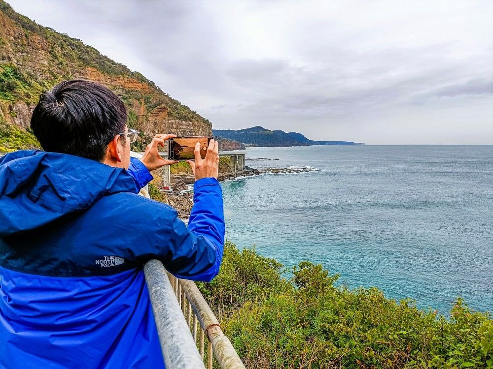 A man in a blue jacket is taking a picture of the ocean.