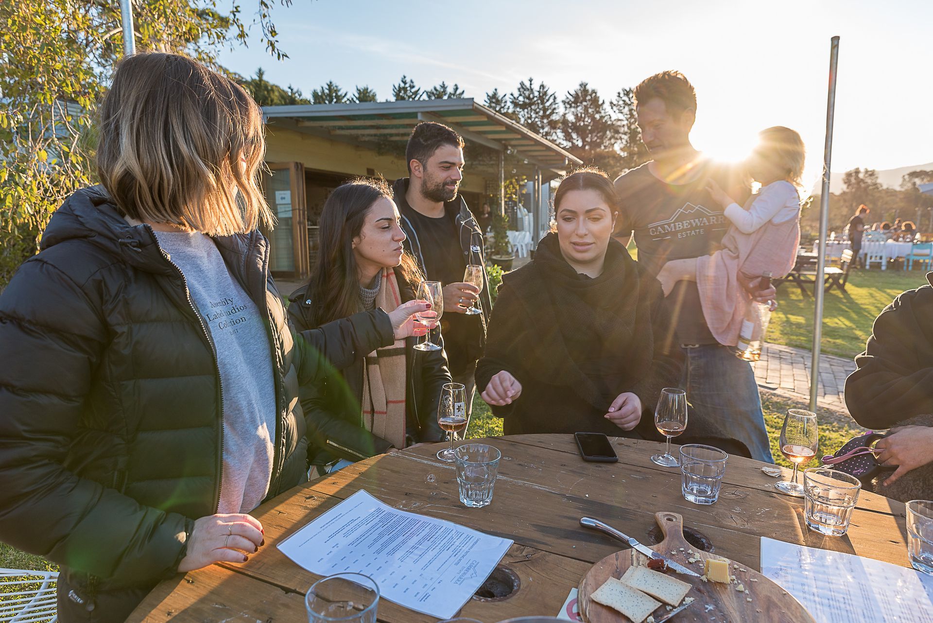 People at a wooden table tasting wine outdoors; sunset in the background.