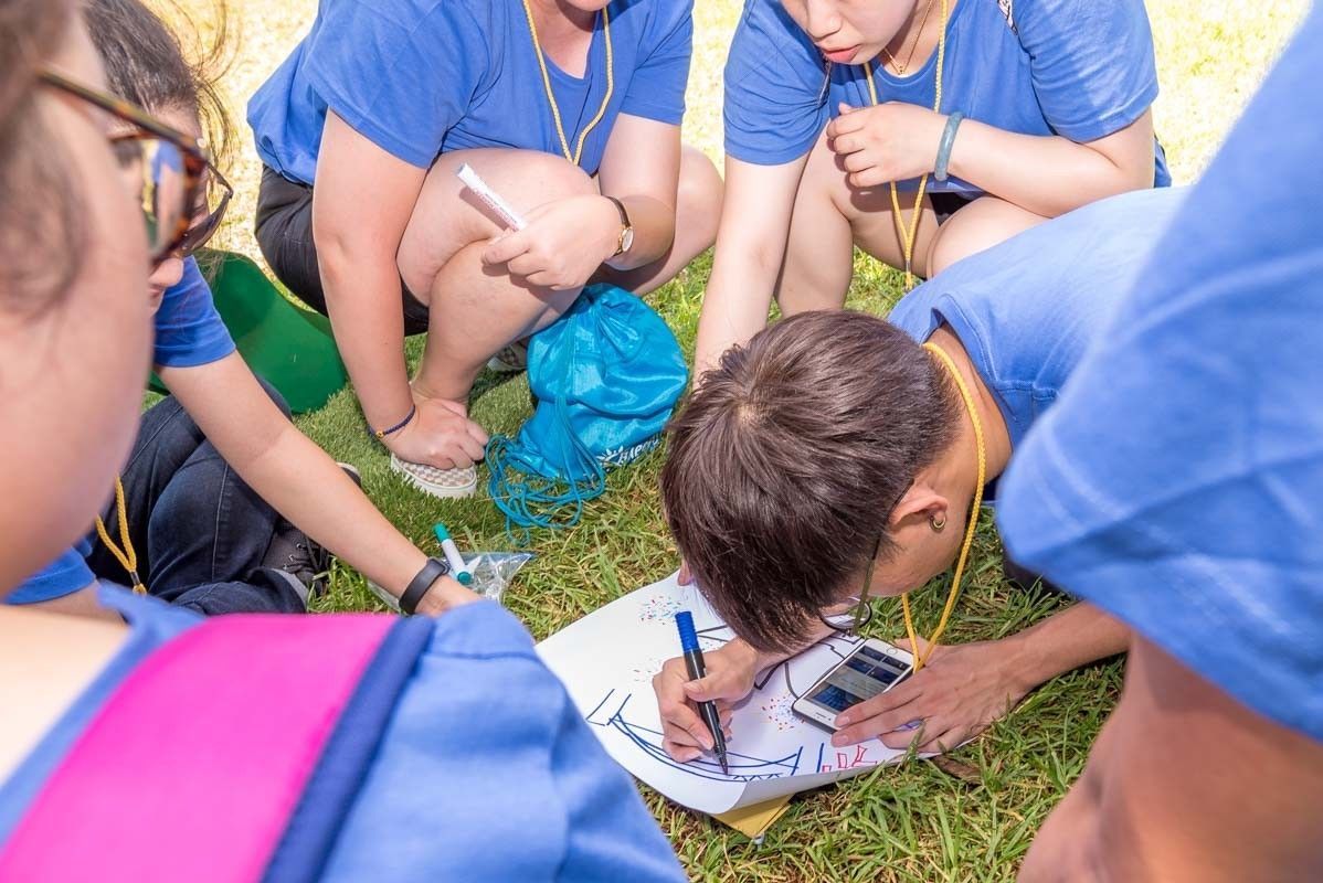 A group in blue shirts huddled on grass, drawing on paper. A person in the center is using a phone and pen.