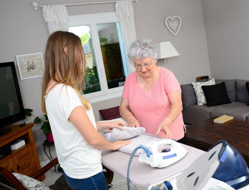 Cheerful Young Girl Ironing And Helping With Household Chores An Elderly Woman At Home — iCan Support Services Kawana QLD