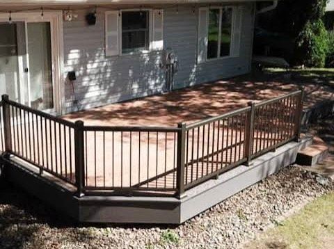 Brown deck with black railing outside a gray house with a sliding door and windows.