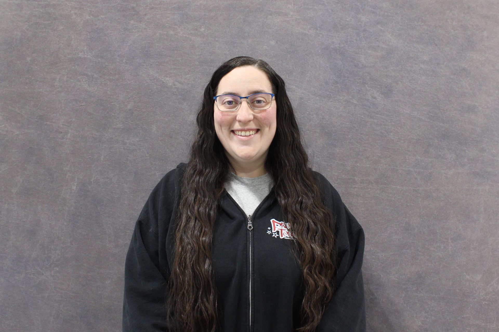 Woman with glasses smiles at the camera, dark hair and black jacket. Gray backdrop.
