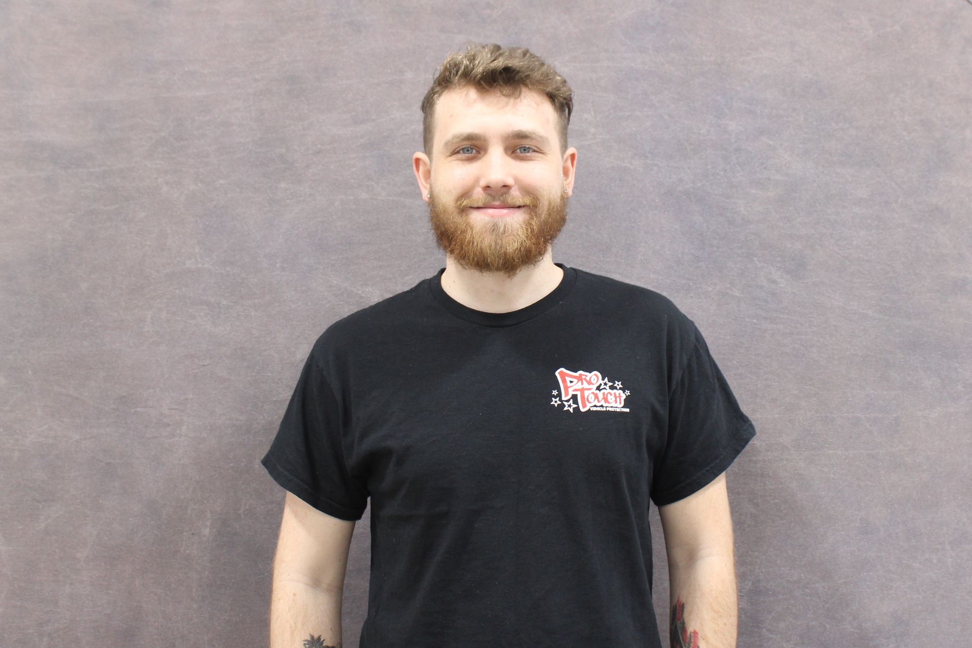Man with a beard, wearing a black t-shirt with a logo, smiling, standing in front of a gray backdrop.