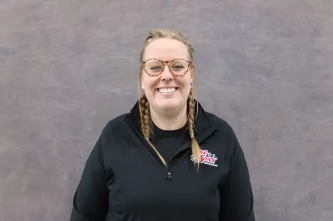 Woman with glasses smiles wearing a black shirt with logo, braids, standing in front of a gray backdrop.