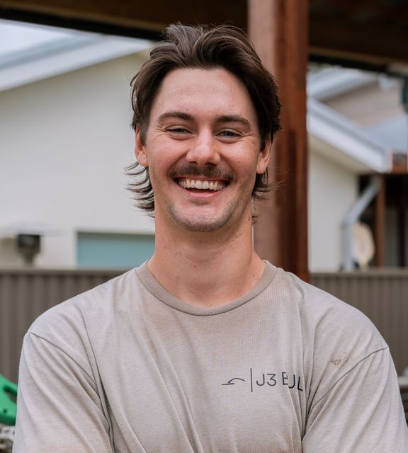 Man with dark hair smiling, wearing a light gray t-shirt. Outdoors, background of house.