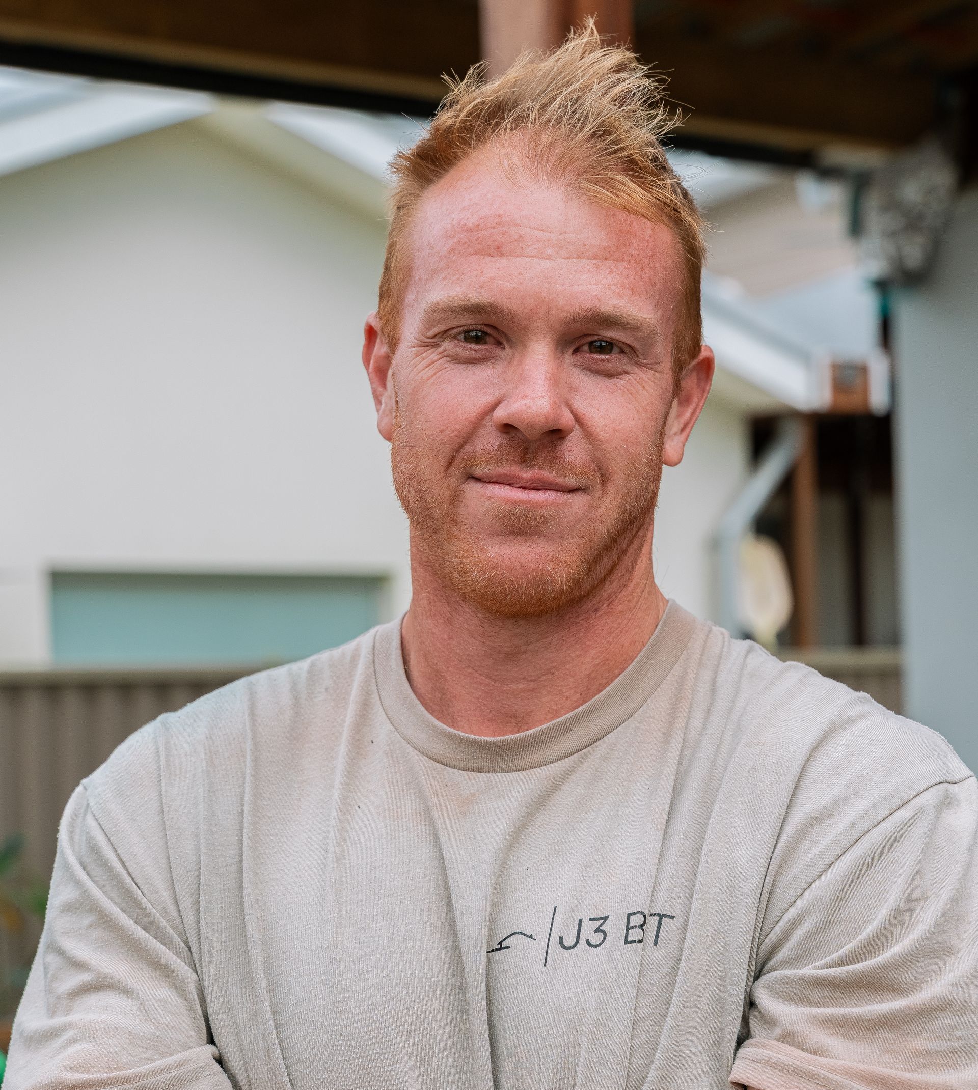 Man with orange-red spiky hair, wearing a beige shirt, smiles outdoors.