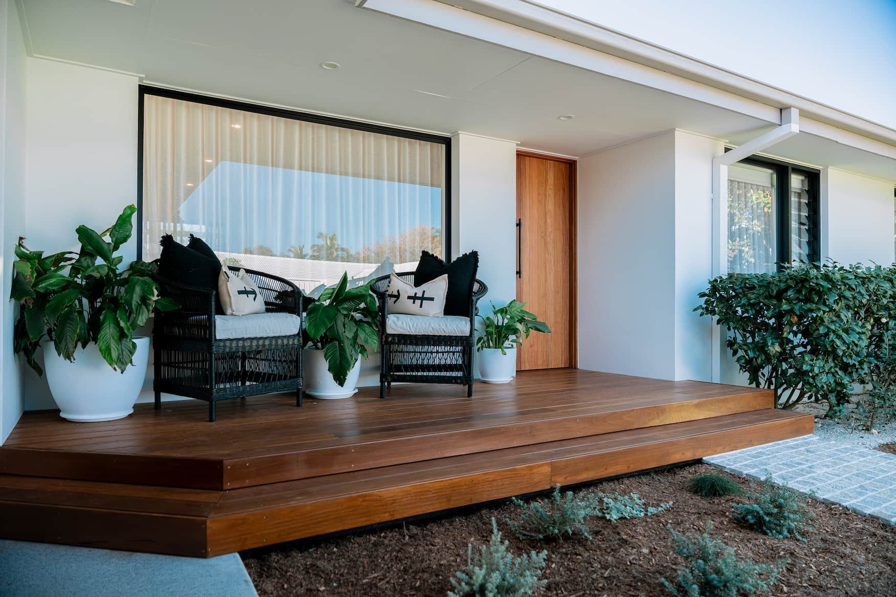 Exterior porch with wooden deck, chairs with pillows, plants in white pots, and a large window.