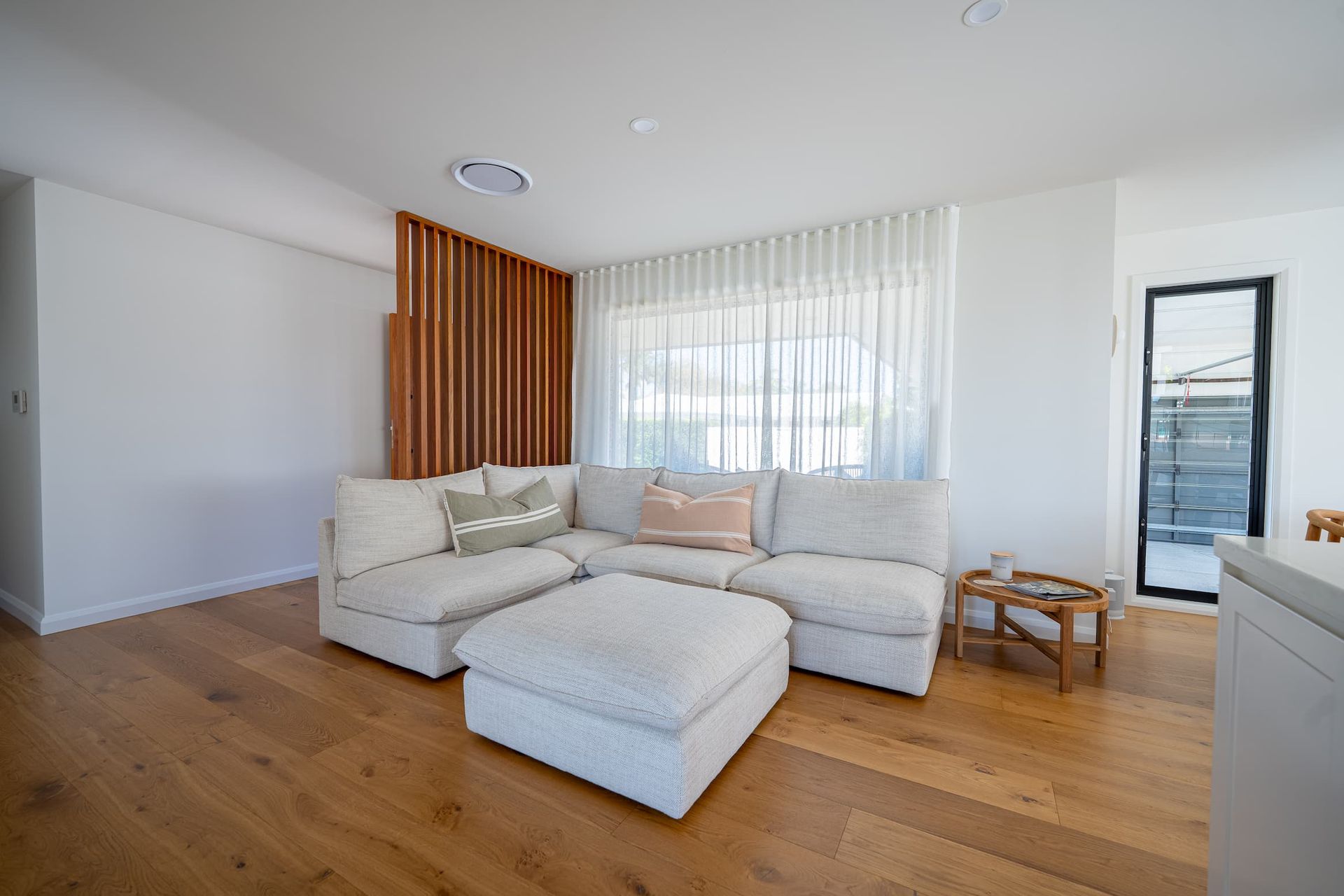 Living room with sectional sofa, wood flooring, wooden slat divider, and large windows with sheer curtains.