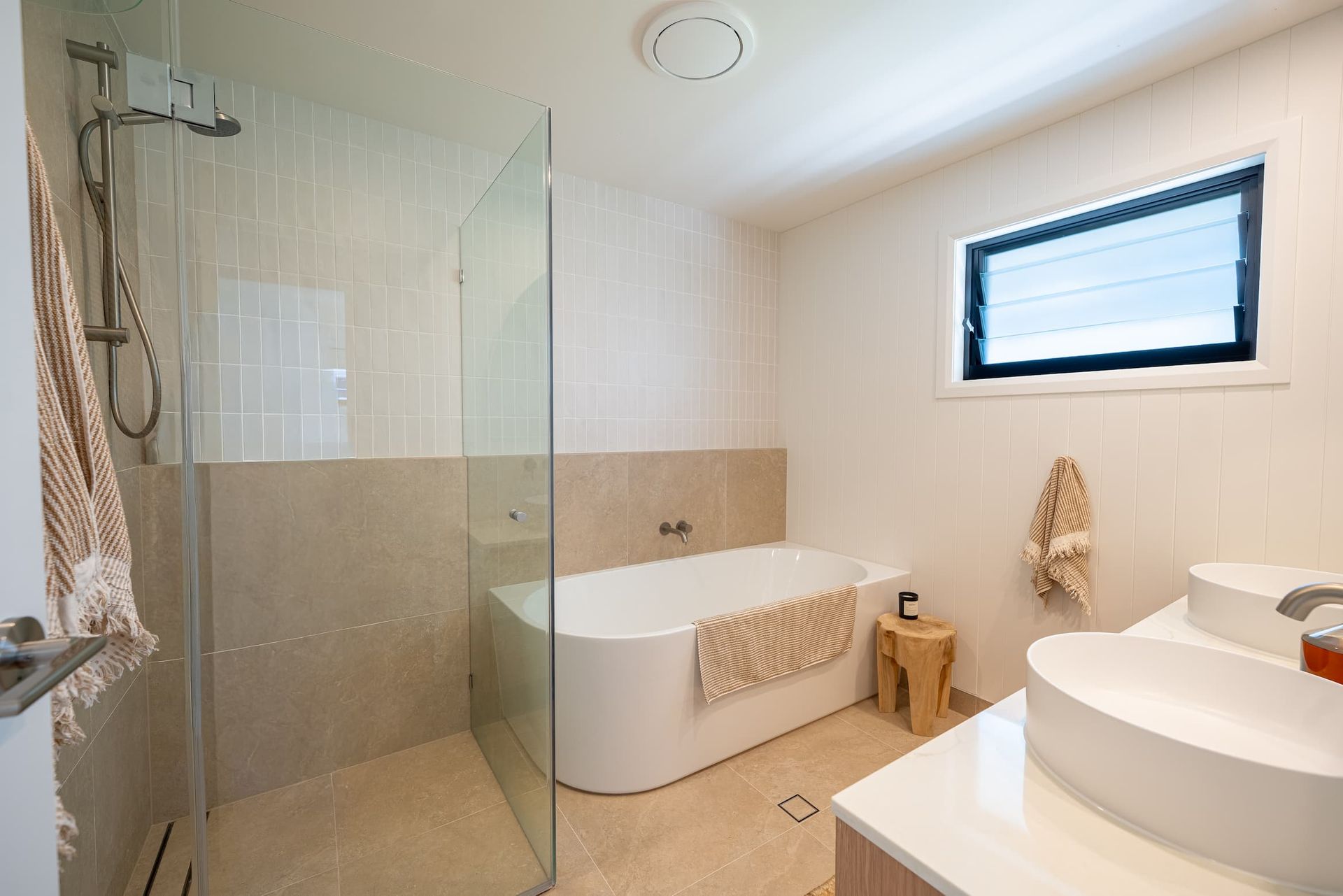 Modern bathroom with a white tub, shower, and sink. Beige tile and white walls. Small wooden stool.