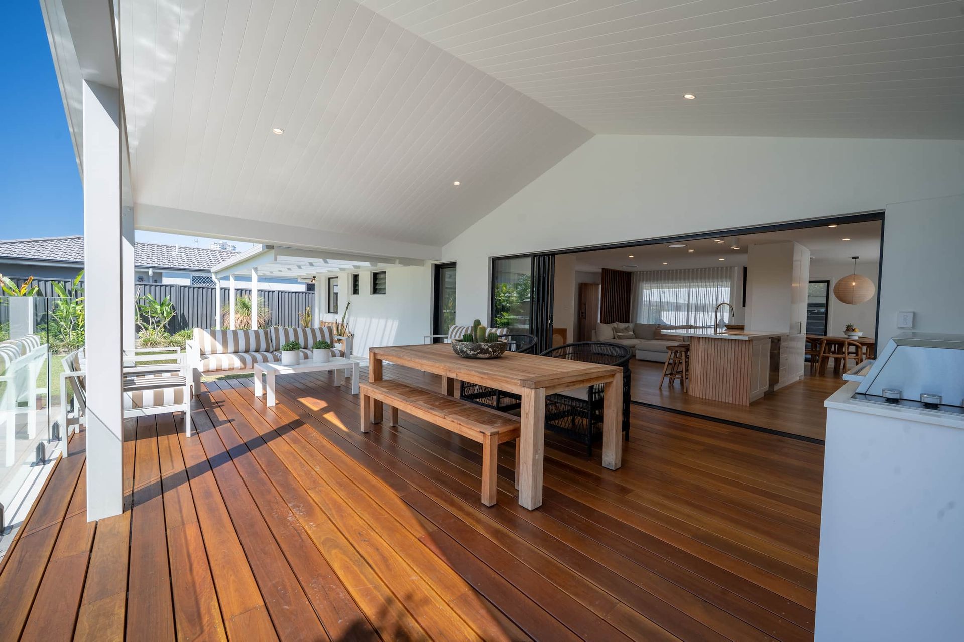 Covered wooden deck with dining table and bench; opens to an indoor kitchen.