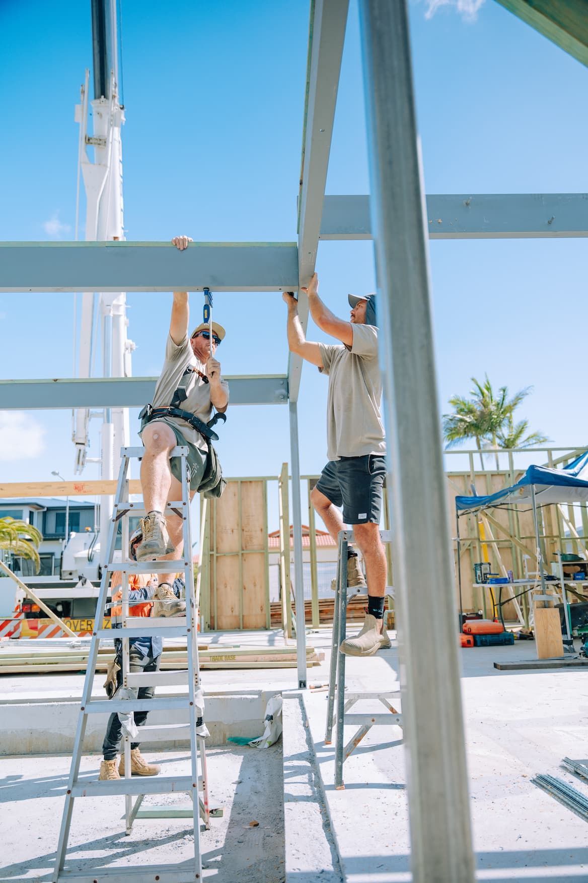 Two construction workers on ladders, attaching metal beams at a construction site on a sunny day.