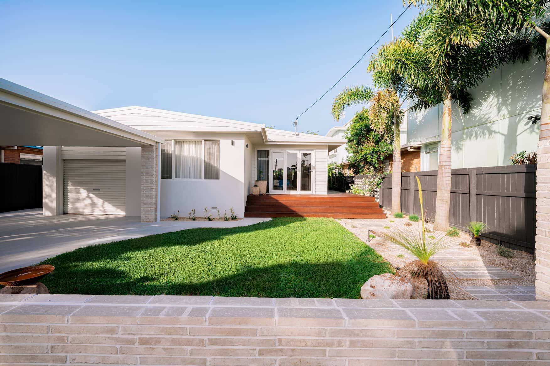 White house with a carport, lawn, and wooden deck. Palm trees and a stone wall are in the yard.