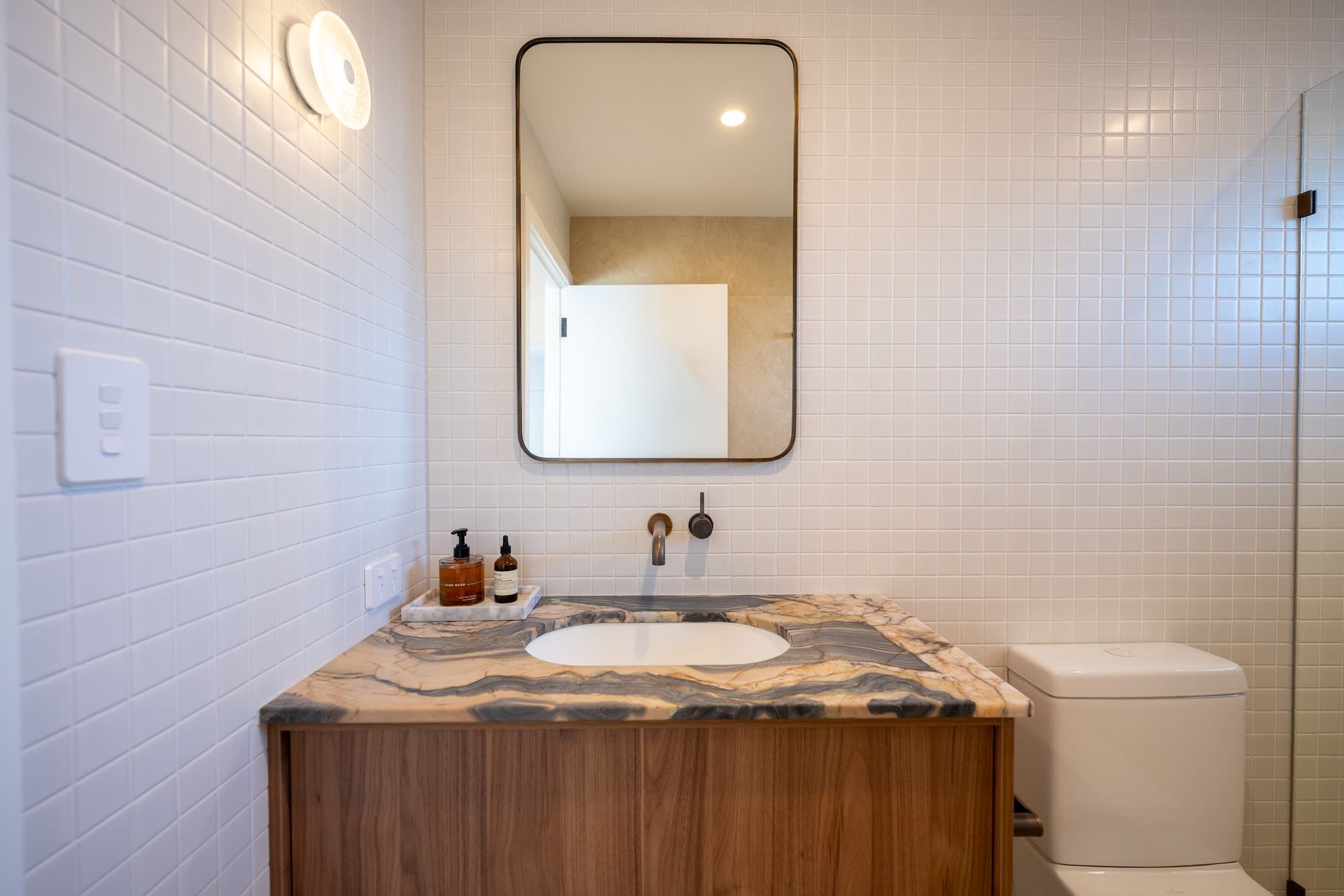 Bathroom with a wooden vanity, marble countertop, and rectangular mirror. White tiled walls, toilet, and glass shower.