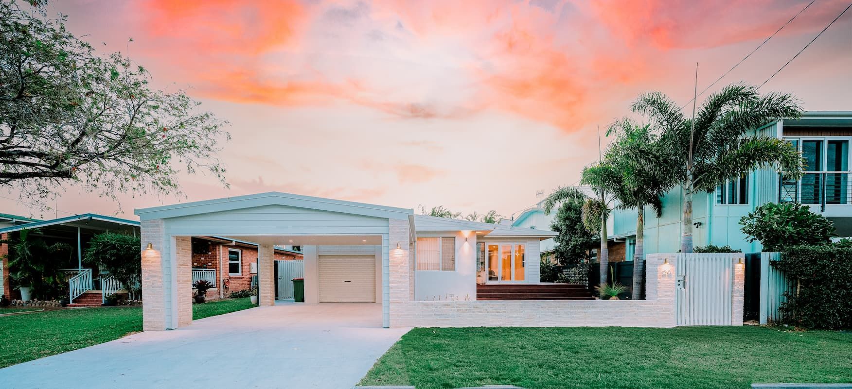 A modern white house with a carport and green lawn against a colorful sky.