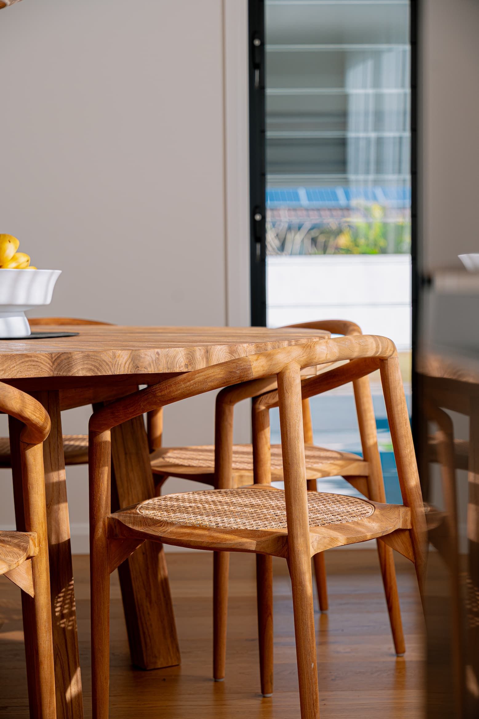 Wooden dining table with woven chairs near a glass door, bathed in sunlight.