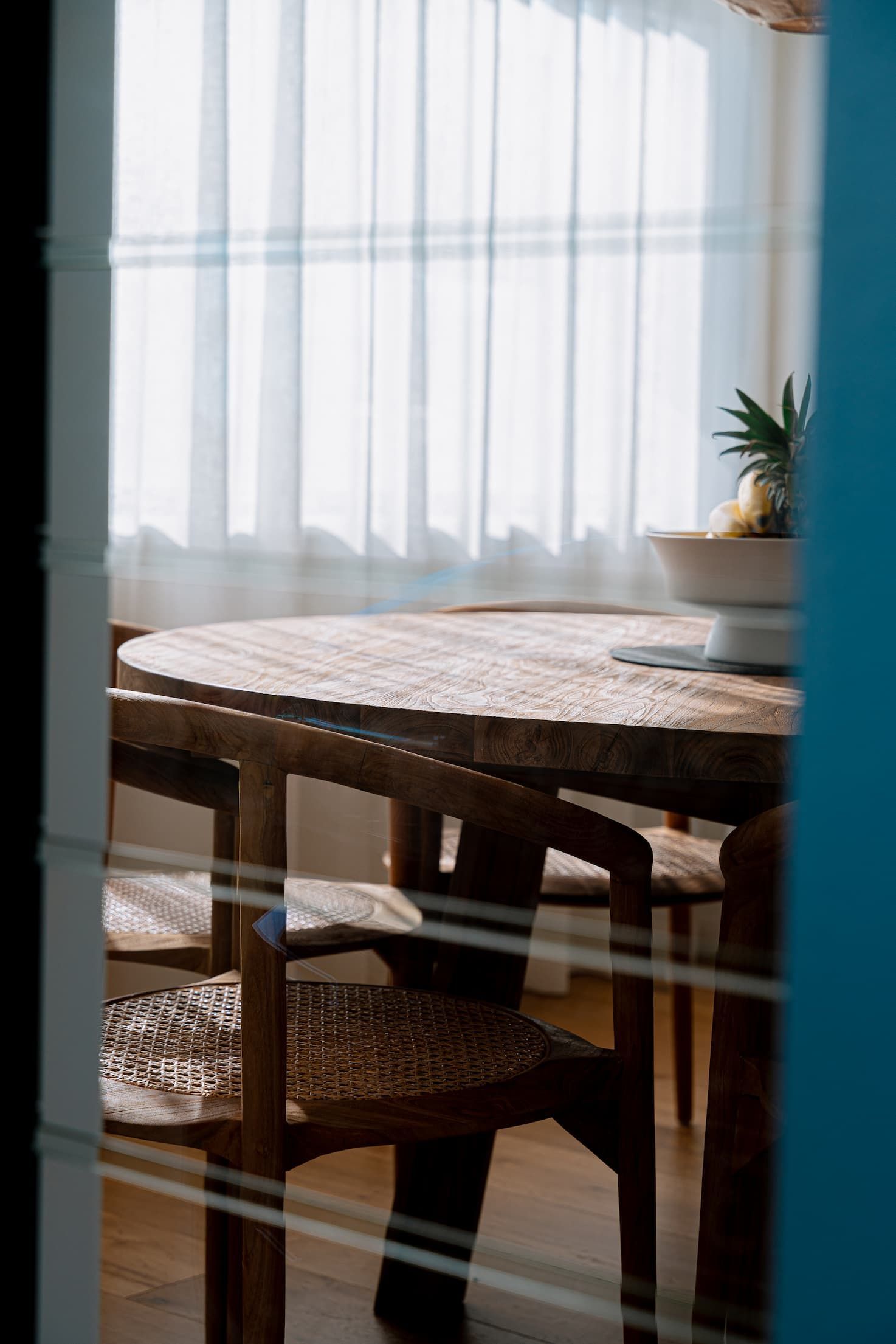 Wooden dining table and chairs near window with sheer curtains. Pineapple in a bowl.