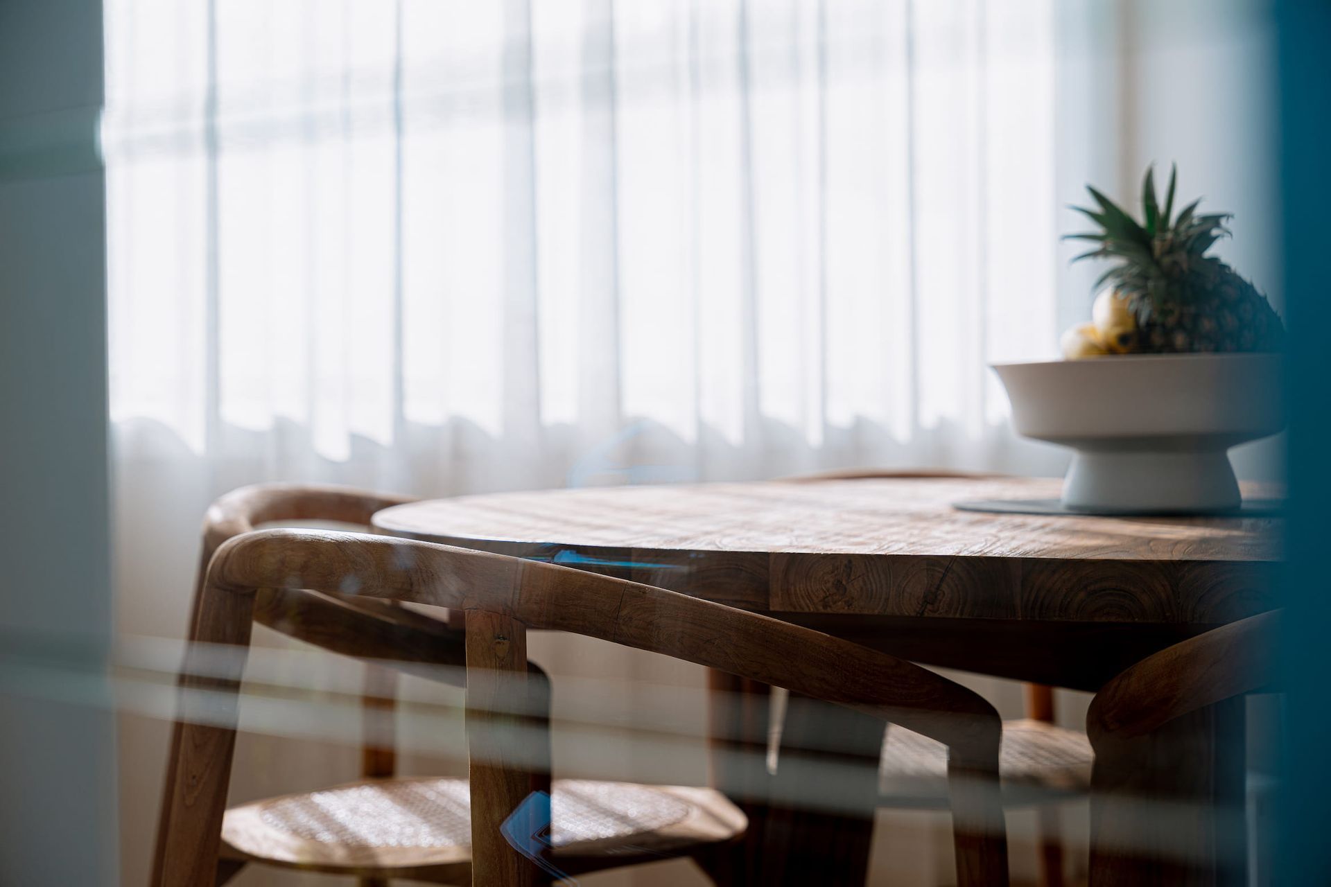 Wooden dining table with chairs, bowl of fruit, sheer curtains.