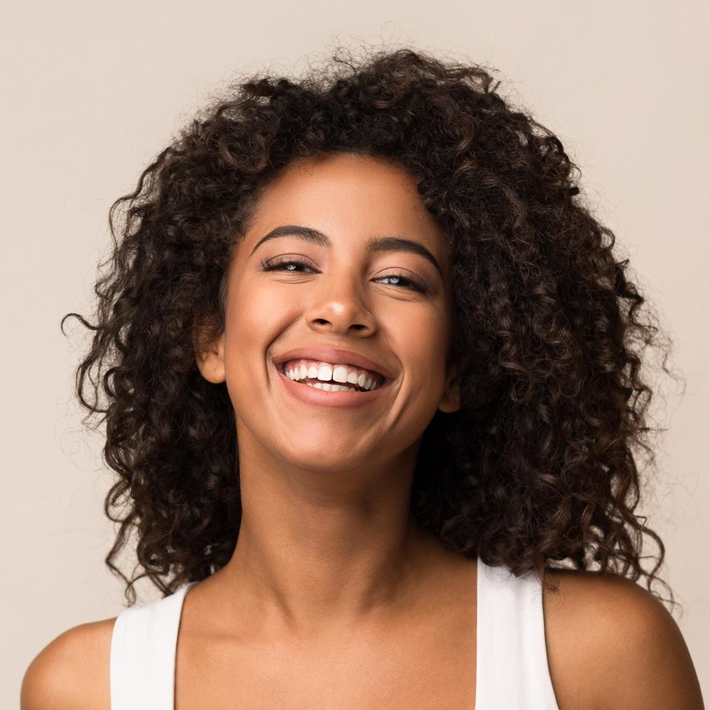 A woman with curly hair is smiling and wearing a white tank top.