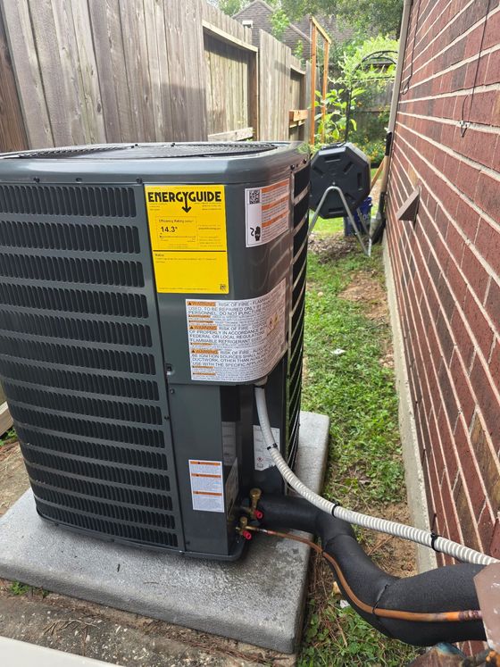 A grey outdoor air conditioning unit sits on a concrete slab next to a brick wall in a grassy backyard.