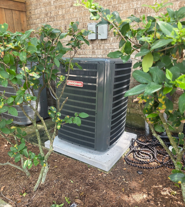 A gray Goodman central air conditioning unit sits on a concrete pad outside a brick house, surrounded by green bushes.