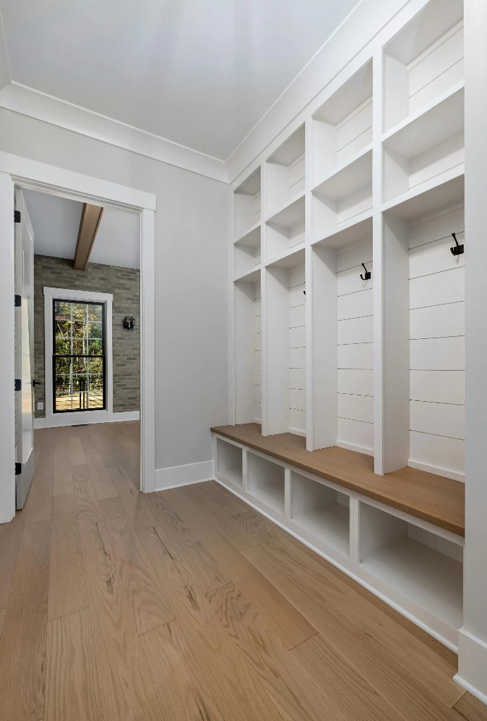 Hallway with white built-in lockers, a wood bench, and light wood flooring. A doorframe leads to a room with a window.