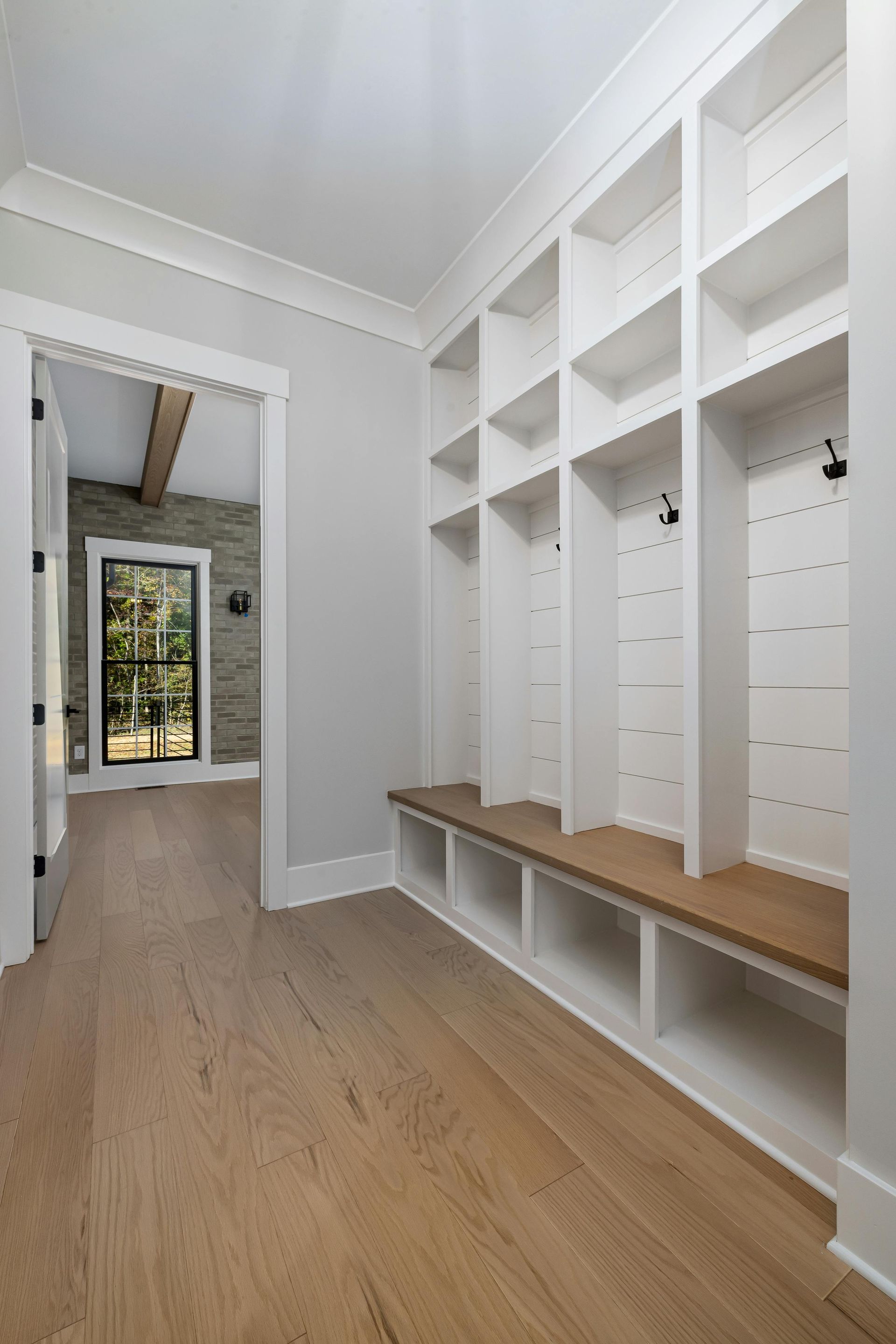 Hallway with white built-in lockers, a wood bench, and light wood flooring. A doorframe leads to a room with a window.