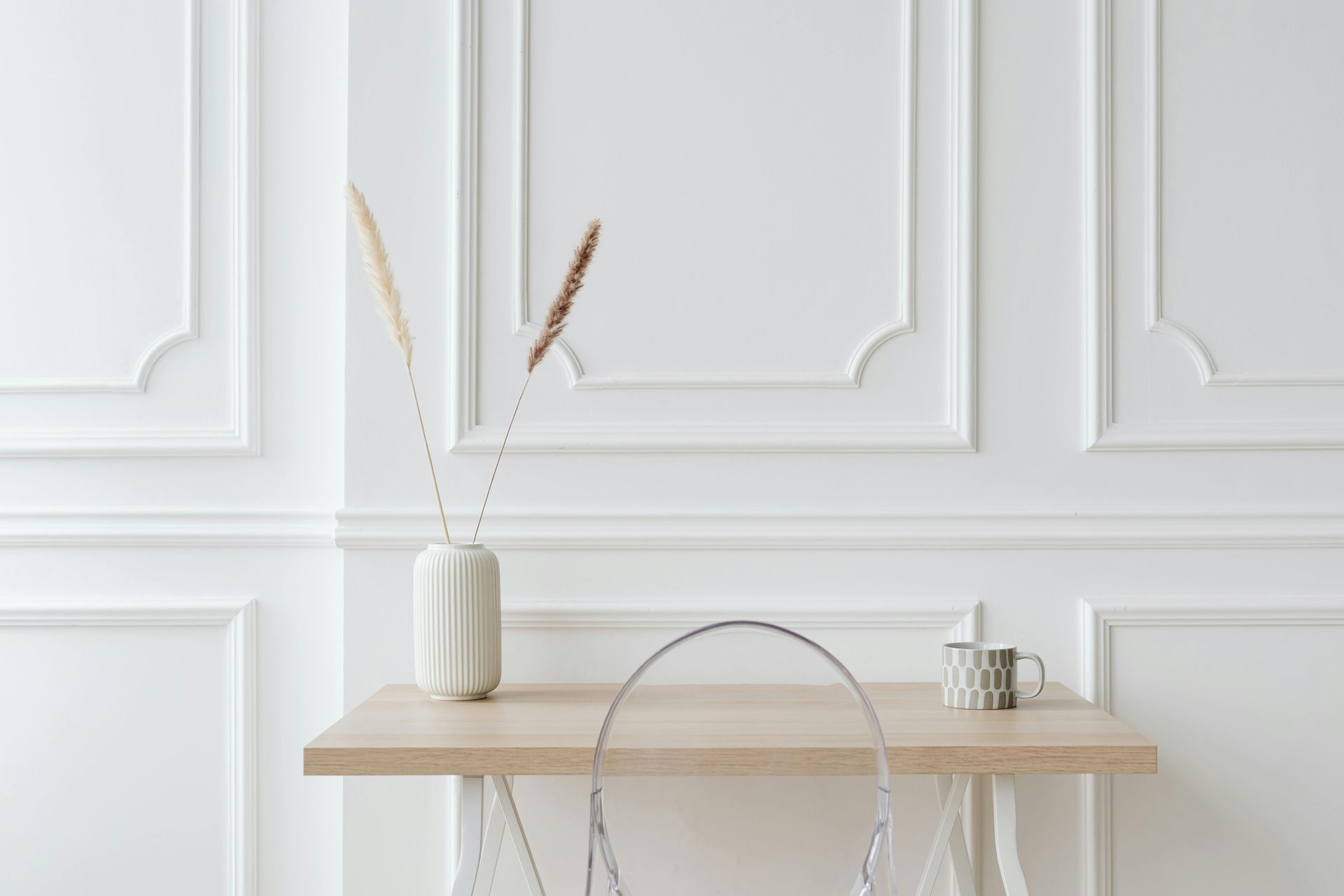 White desk with a transparent chair, vase with dried grass, and a mug against a white paneled wall.