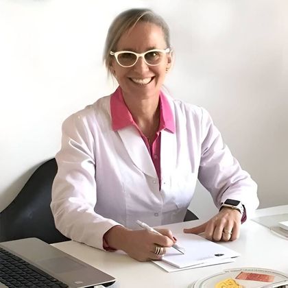 Una mujer sonriente con una bata blanca, gafas y una camisa rosa está sentada en un escritorio, sosteniendo un bolígrafo y mirando papeles.