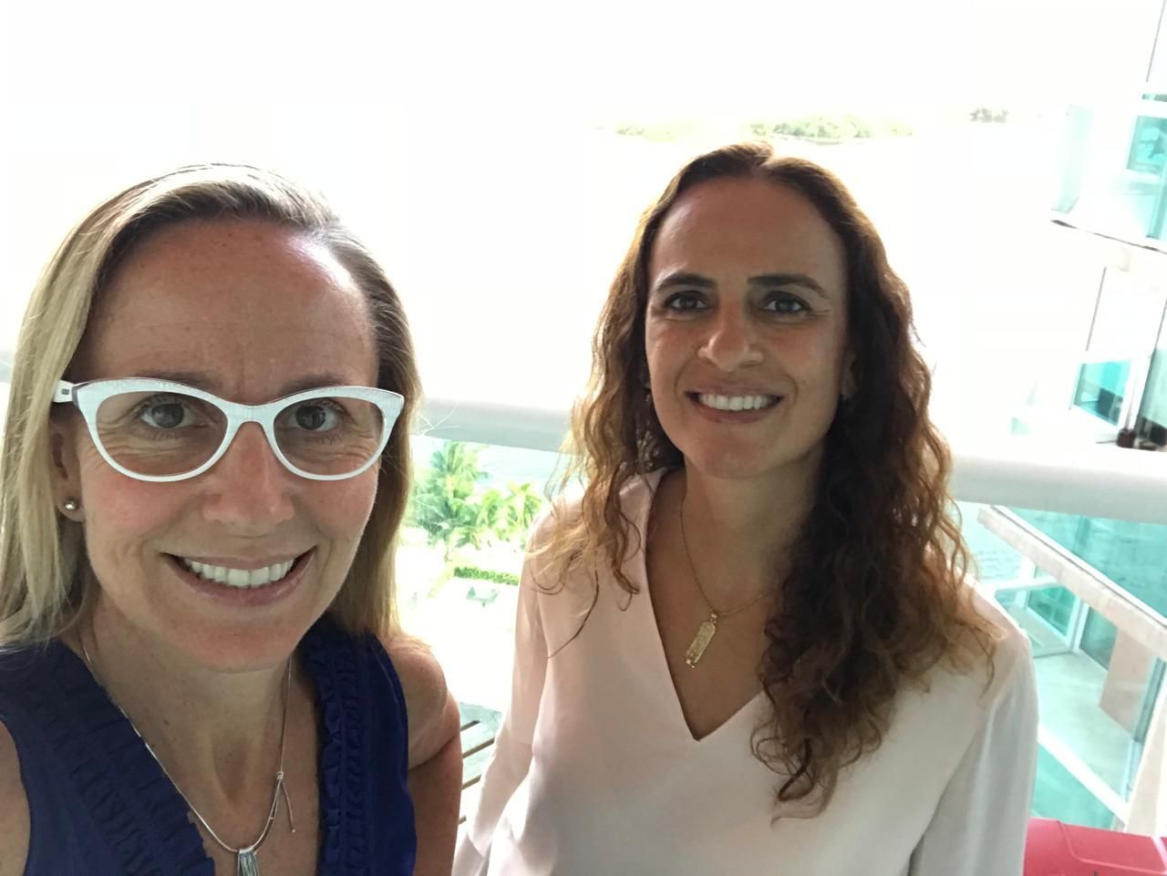 Dos mujeres sonriendo en un balcón. Una lleva gafas, la otra, un collar. Vistas al agua.