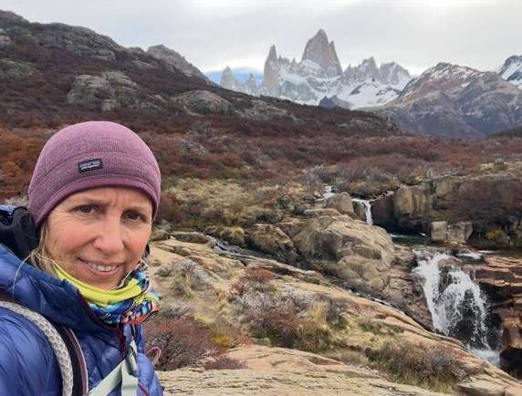 Una mujer sonríe a la cámara cerca de una cascada y unas montañas, vistiendo sombrero y chaqueta.