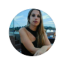 Mujer con cabello largo y rubio, con un vestido negro, sentada afuera con un fondo borroso de agua y cielo.