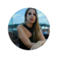 Mujer con cabello largo y rubio, con un vestido negro, sentada afuera con un fondo borroso de agua y cielo.