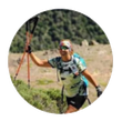Una mujer corre cuesta arriba con bastones de senderismo. Lleva gafas de sol, una mochila de hidratación y pantalones cortos negros, con las montañas de fondo.