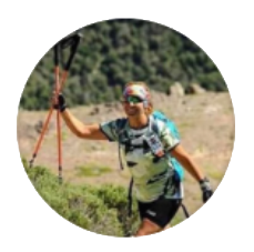 Una mujer corre cuesta arriba con bastones de senderismo. Lleva gafas de sol, una mochila de hidratación y pantalones cortos negros, con las montañas de fondo.