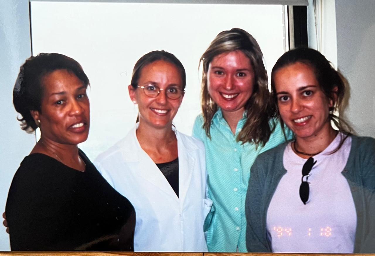Cuatro mujeres sonrientes posando en un interior. Una lleva bata, otra camisa azul y la tercera un cárdigan gris claro.