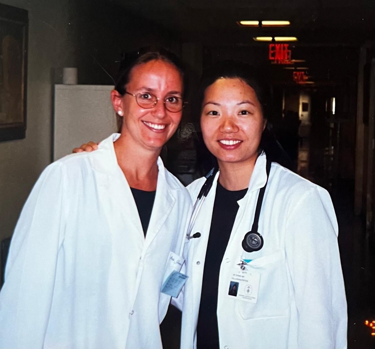 Dos mujeres con batas blancas sonríen en un pasillo de hospital. Una lleva gafas y rodea con el brazo a la otra, que porta un estetoscopio.
