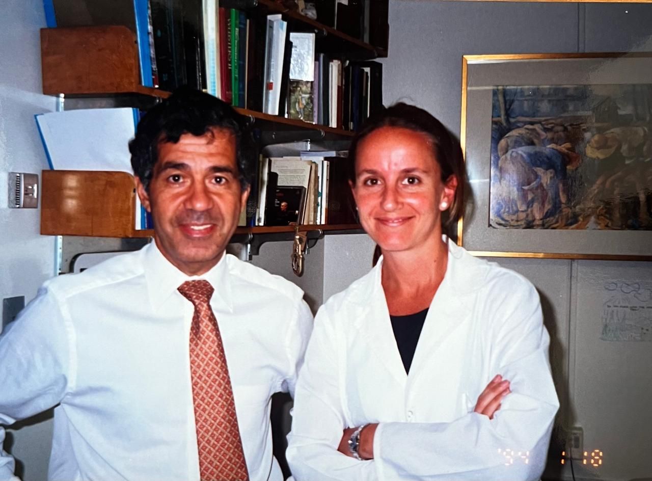 Hombre y mujer de pie juntos en un interior; el hombre sonríe, vistiendo una corbata; la mujer tiene los brazos cruzados, vistiendo una bata de laboratorio.