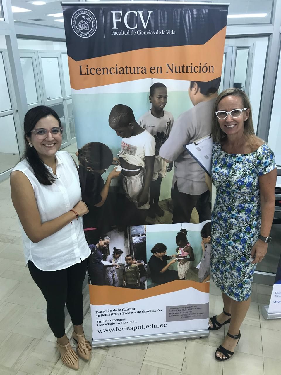 Dos mujeres están junto a una pancarta que anuncia un programa de nutrición. Una sonríe, la otra sostiene un papel.