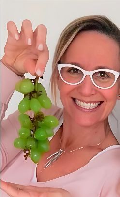 Mujer sosteniendo uvas verdes, sonriendo, usando gafas y un collar.