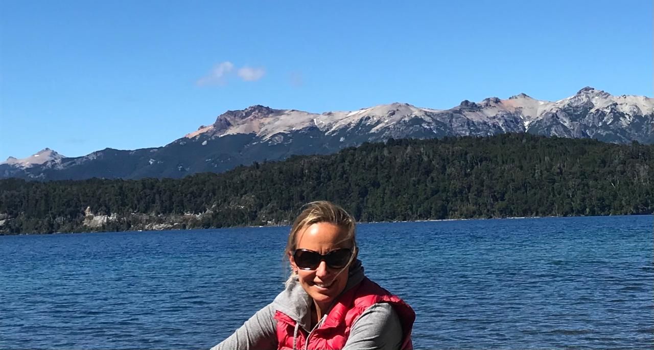 Una mujer con gafas de sol y chaleco se sienta junto a un lago, con montañas al fondo y un cielo azul.