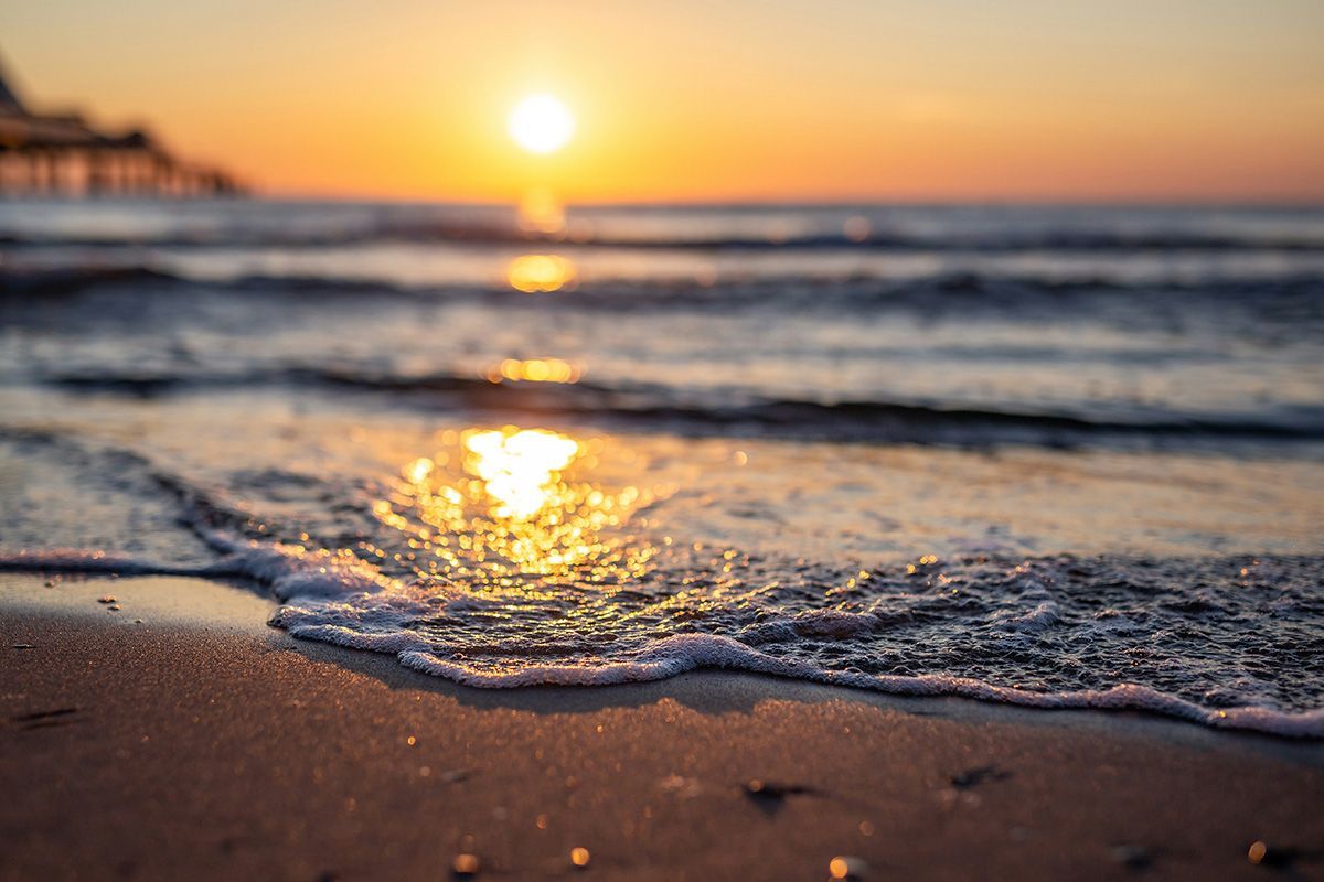 Sunset over ocean with waves gently washing ashore; bright orange sun reflected on the water.