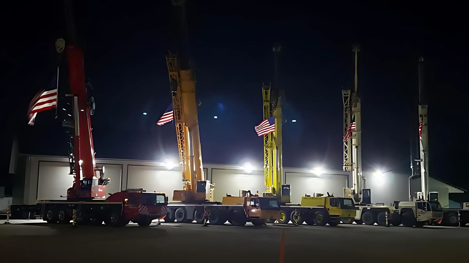 A row of construction vehicles are parked in a parking lot at night
