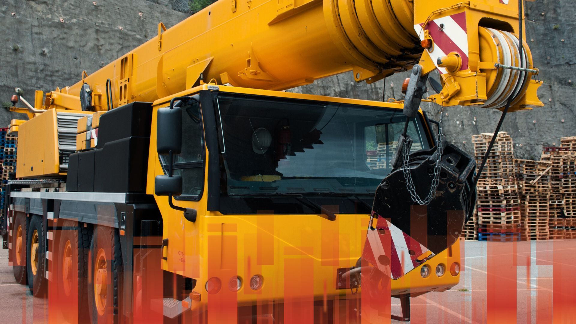 A large yellow crane is parked in front of a pile of wooden pallets.