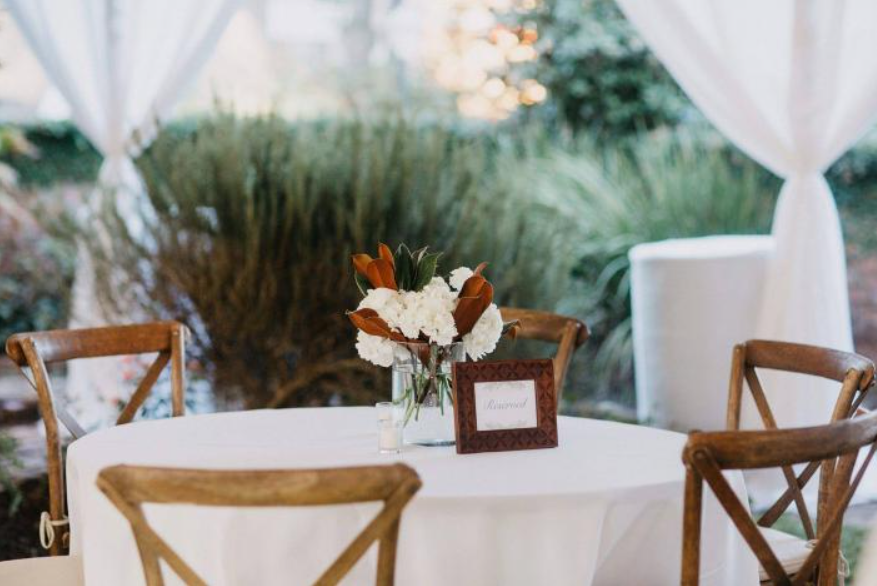 Round white table with tan chairs and a floral centerpiece under a white tent outdoors