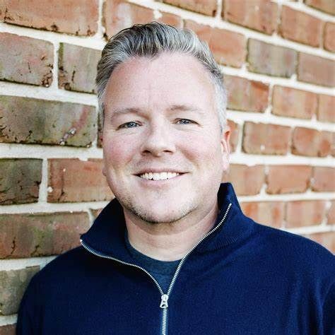 A man in a blue sweater is smiling in front of a brick wall.