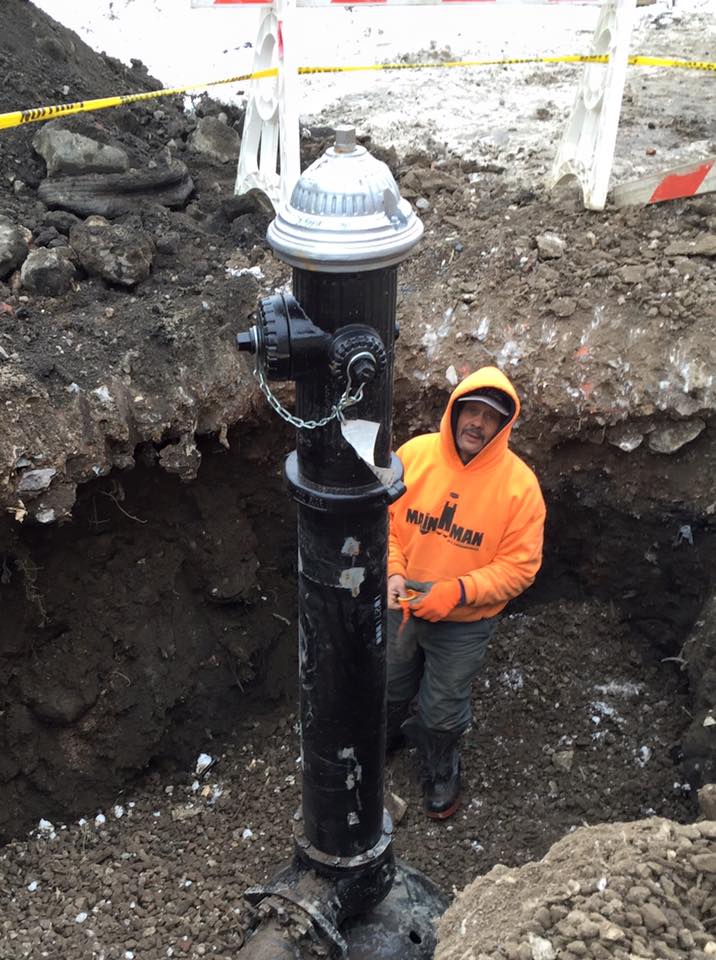 A man in an orange hoodie is standing next to a fire hydrant in the dirt.
