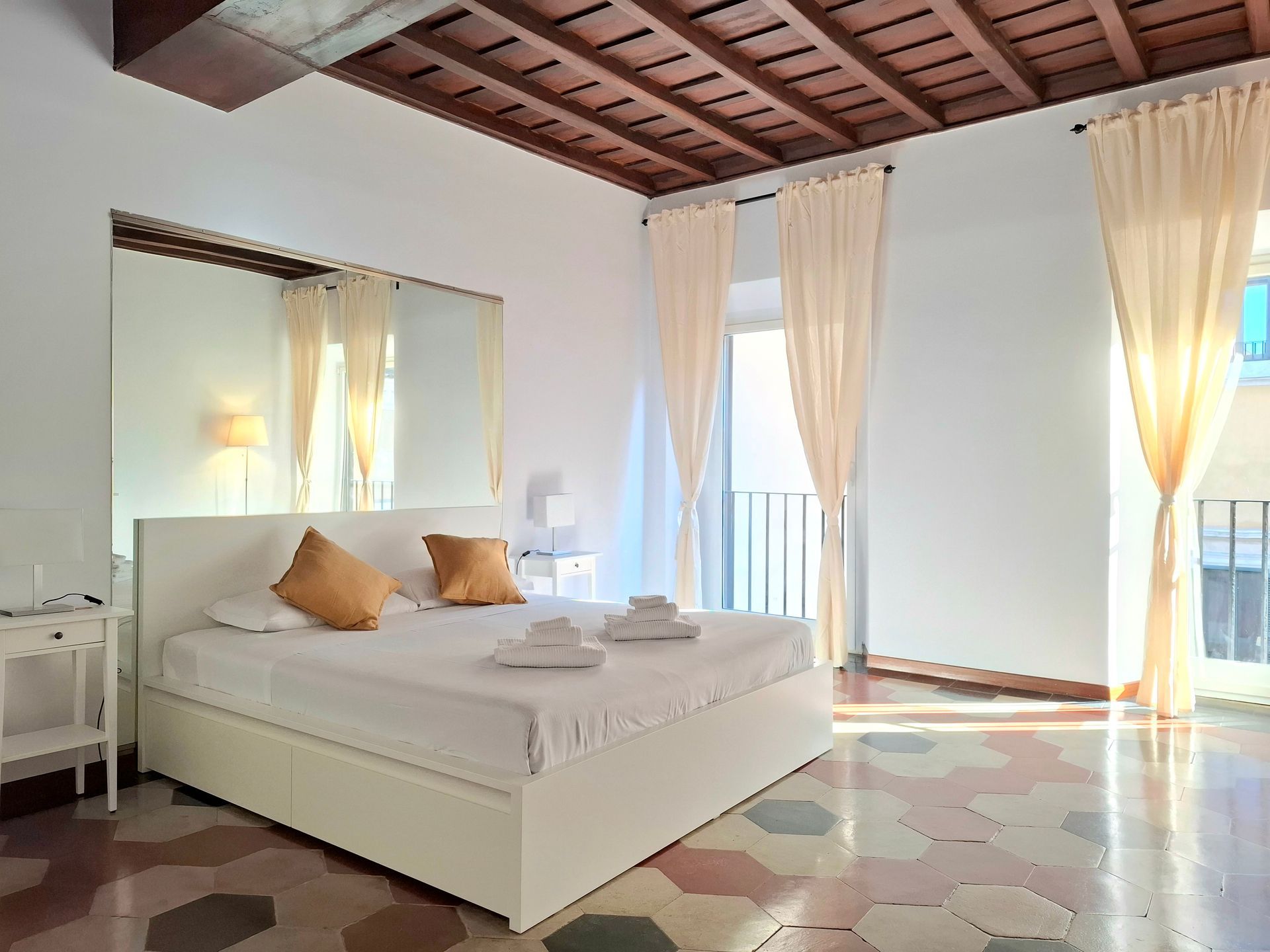 Camera da letto con letto bianco, tende beige, balcone e pavimento in piastrelle geometriche.