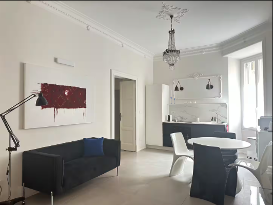 Soggiorno con divano nero, opere d'arte astratte, cucina, tavolo da pranzo e lampadario.